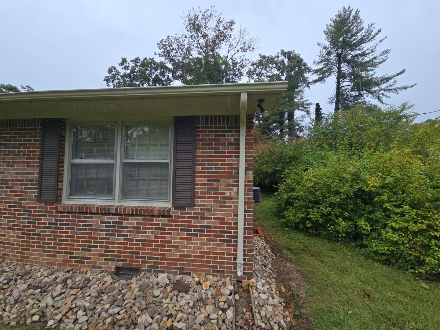Red brick house with window, brown shutters, and green bushes on a hillside.