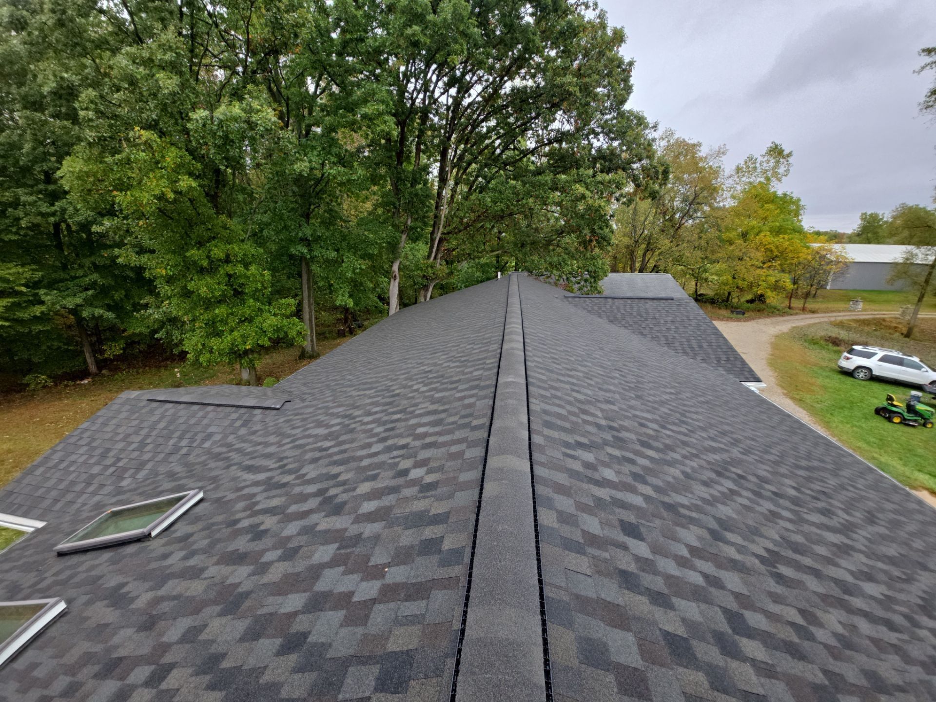 Dark gray asphalt shingle roof, surrounded by green trees and a cloudy sky.