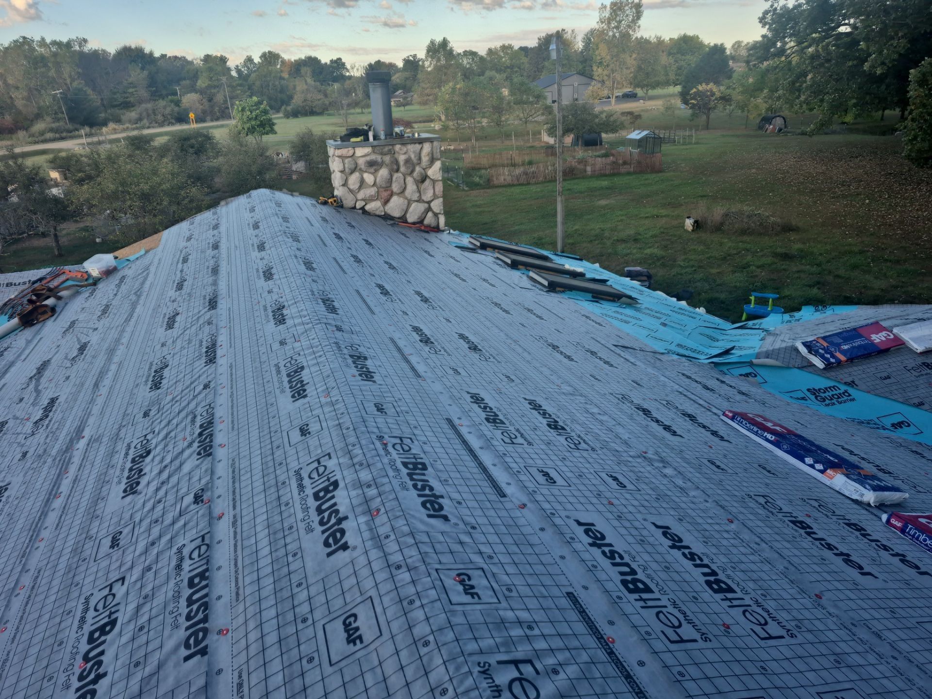 Rooftop partially covered with black underlayment, stone chimney, green field in background.