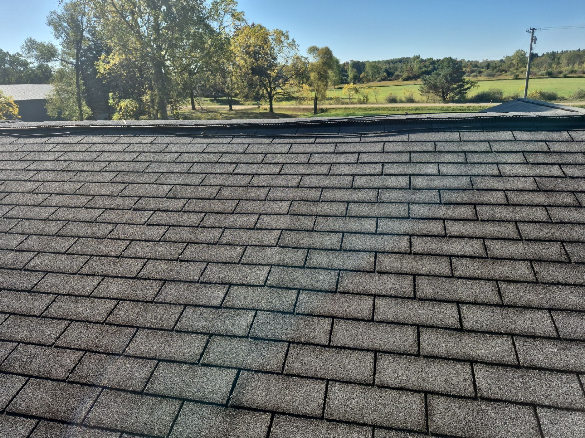 Gray asphalt shingle roof, outdoor setting with trees and field visible in the distance.