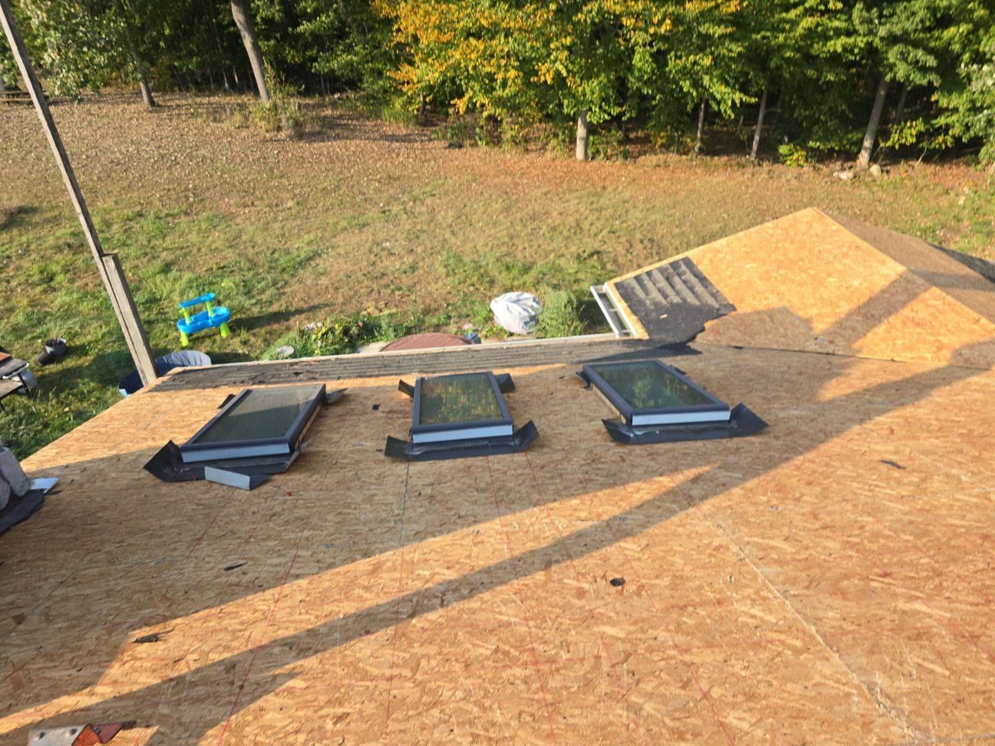 Three skylights installed on a roof covered in wood panels, with a grassy area in the background.