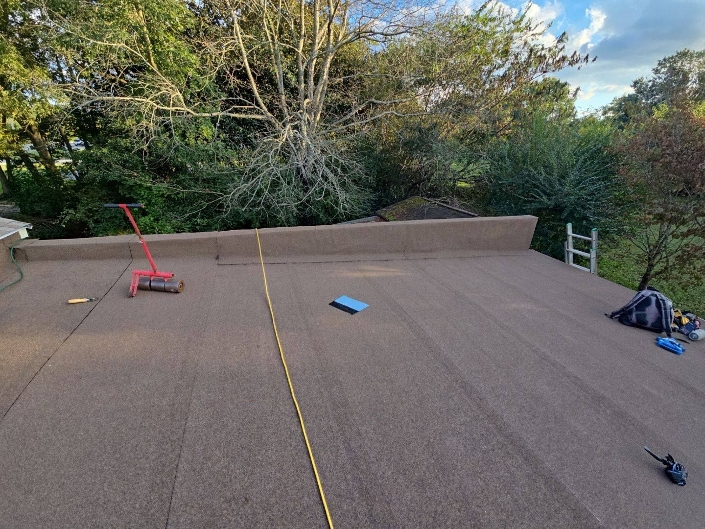 Flat roof with brown surface; gardening tools, rope, and blue pad are visible. Green trees in the background.