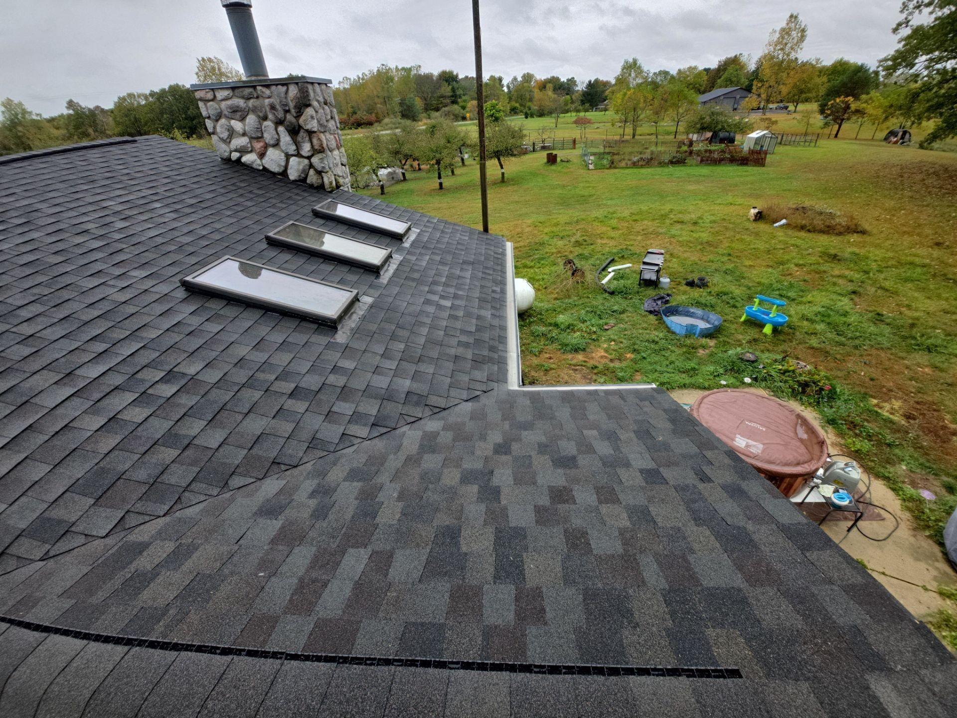 A dark gray shingled roof with three skylights, stone chimney, and a view of a grassy field.