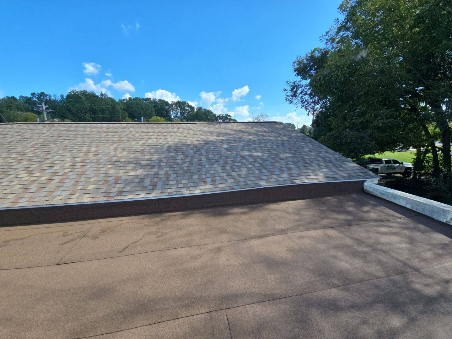 Overhead view of a roof with brown shingles, brown flat surface, and blue sky with clouds in the background.