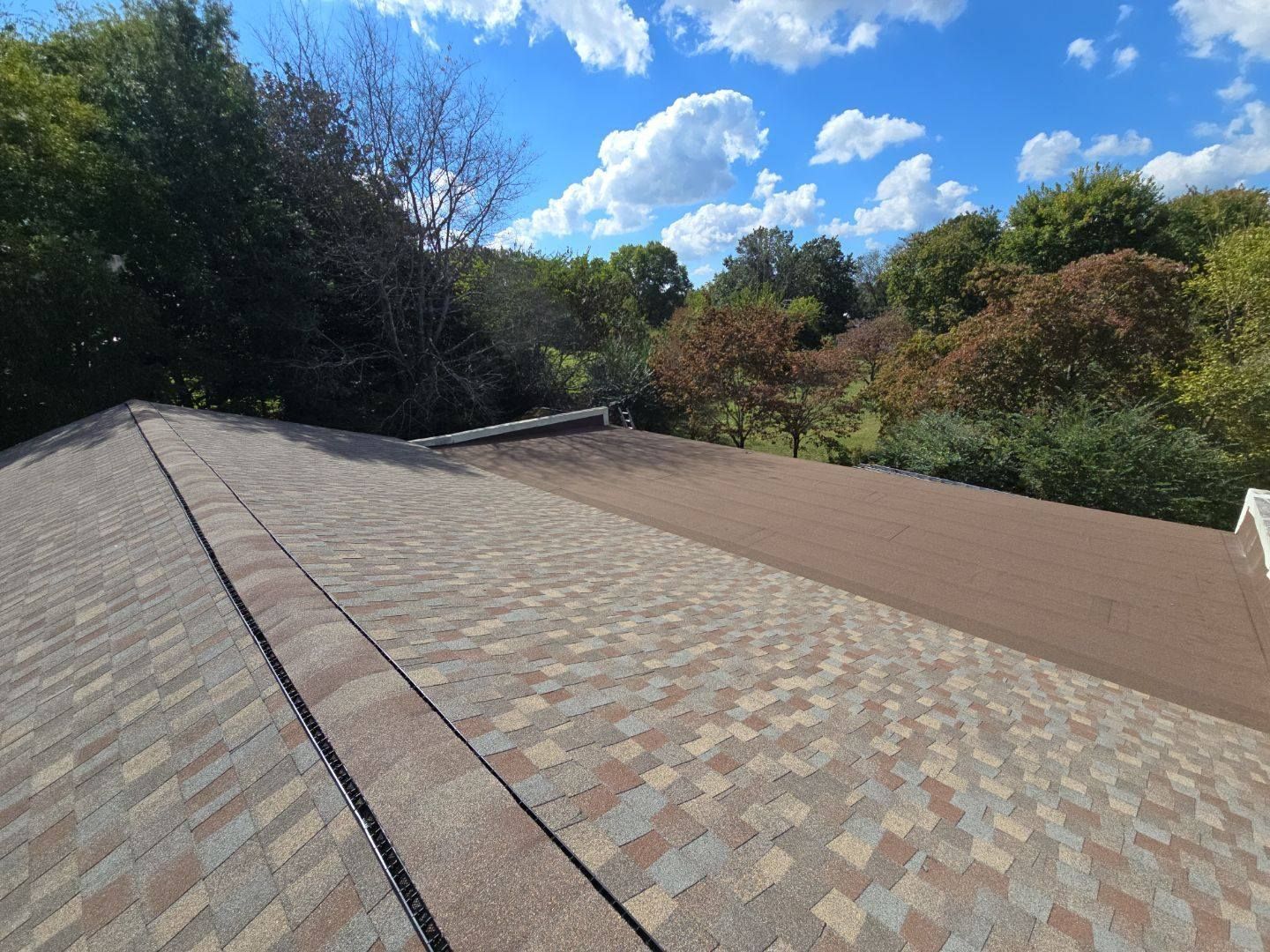 Rooftop with brown and tan shingles, against a backdrop of trees and a blue sky with clouds.