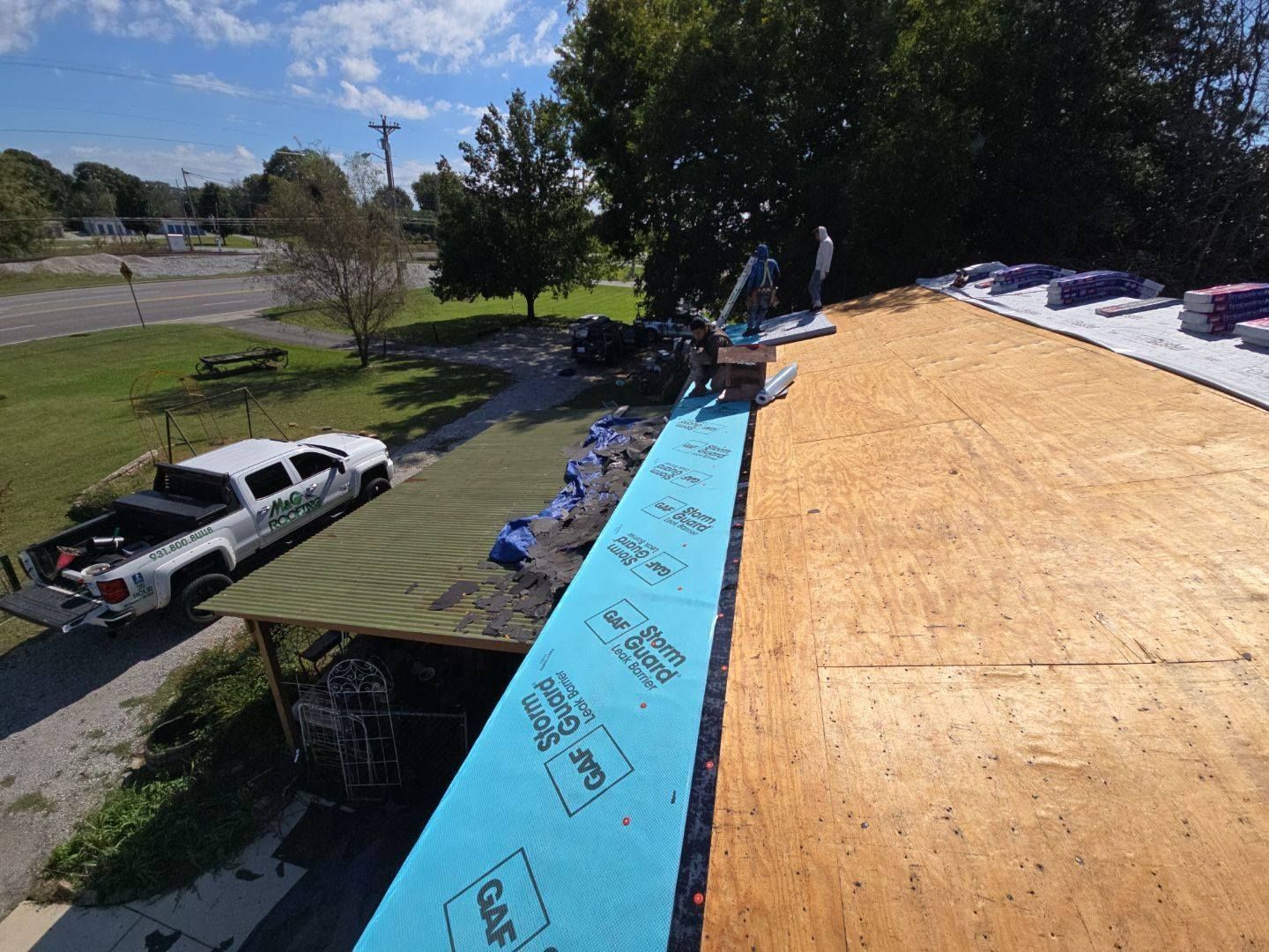 Roofers installing blue underlayment on a wooden roof. A white truck and trees are in the background.