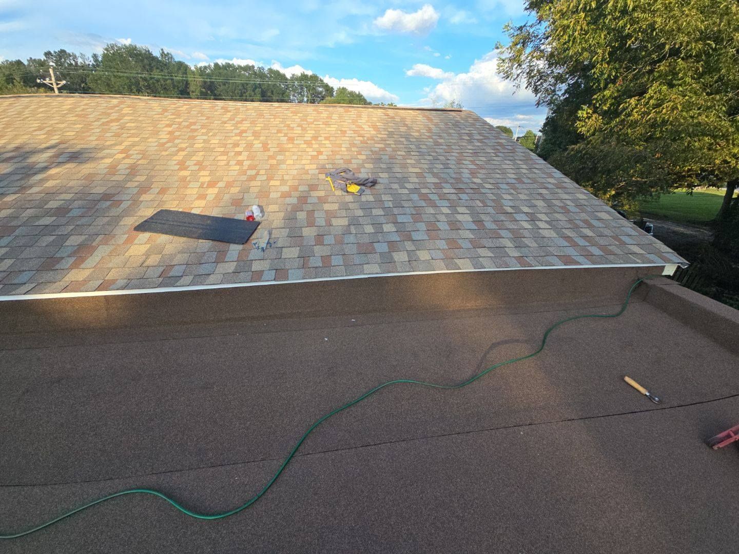 A rooftop with brown shingles and a flat, dark surface, with tools and a green hose present.