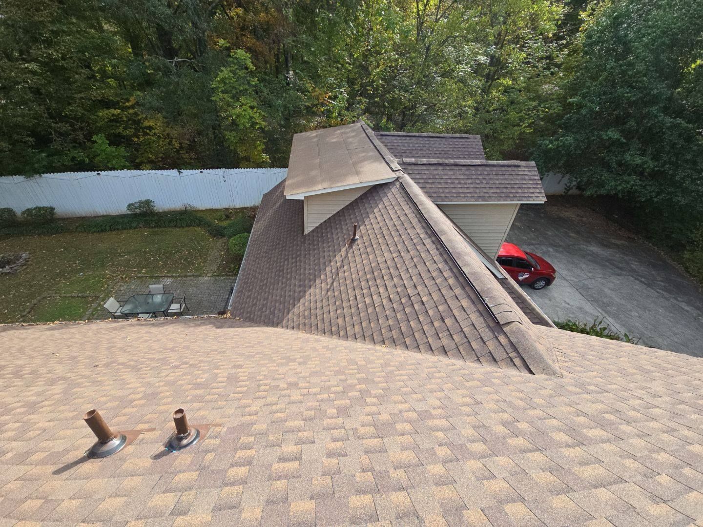 Overhead view of a roof with brown shingles. Trees, white fence, and a red car are in the background.