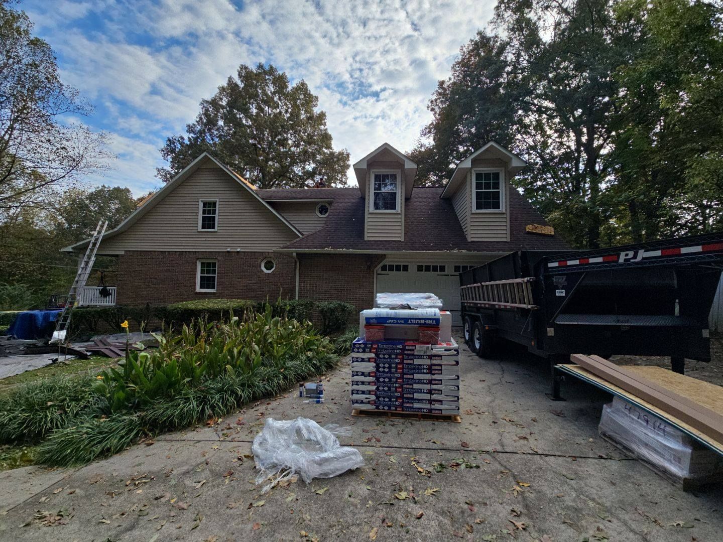 House with roof under construction, pallets of shingles on driveway, trailer and ladder.