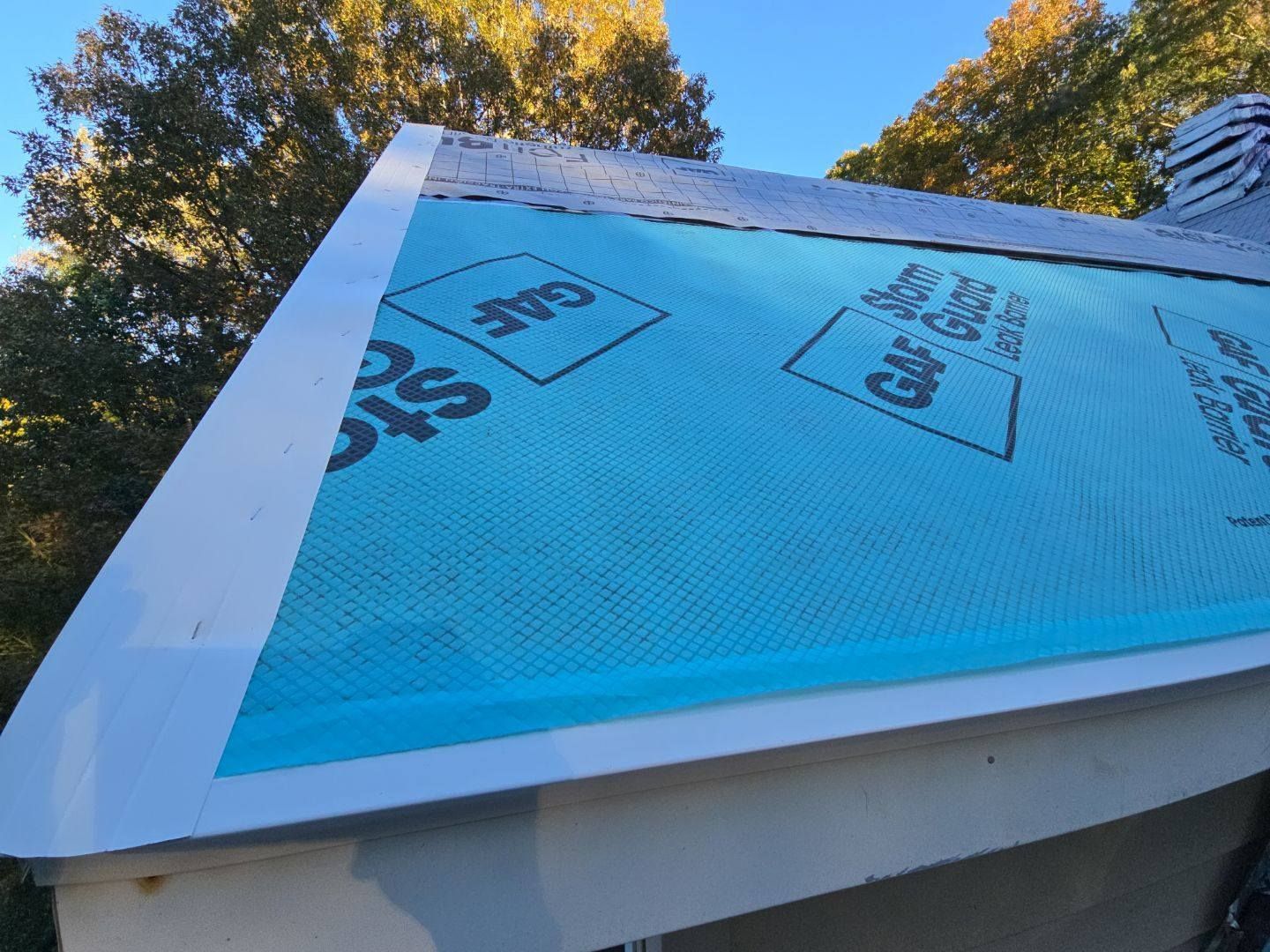 Close-up of a roof edge with white trim and blue underlayment marked with the GAF logo, trees in background.