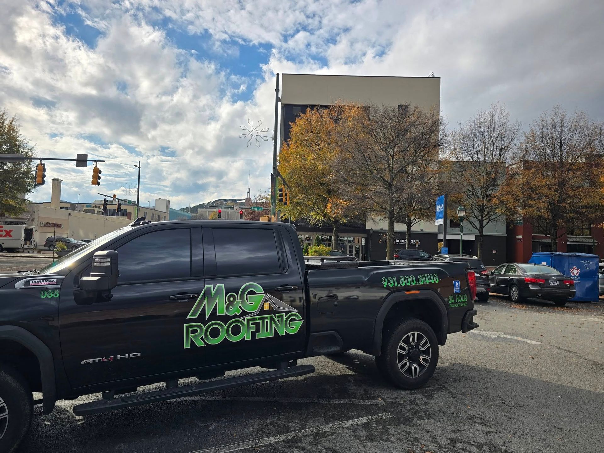 Black M&G Roofing truck parked on a city street. Buildings, trees, and traffic lights in background under cloudy sky.