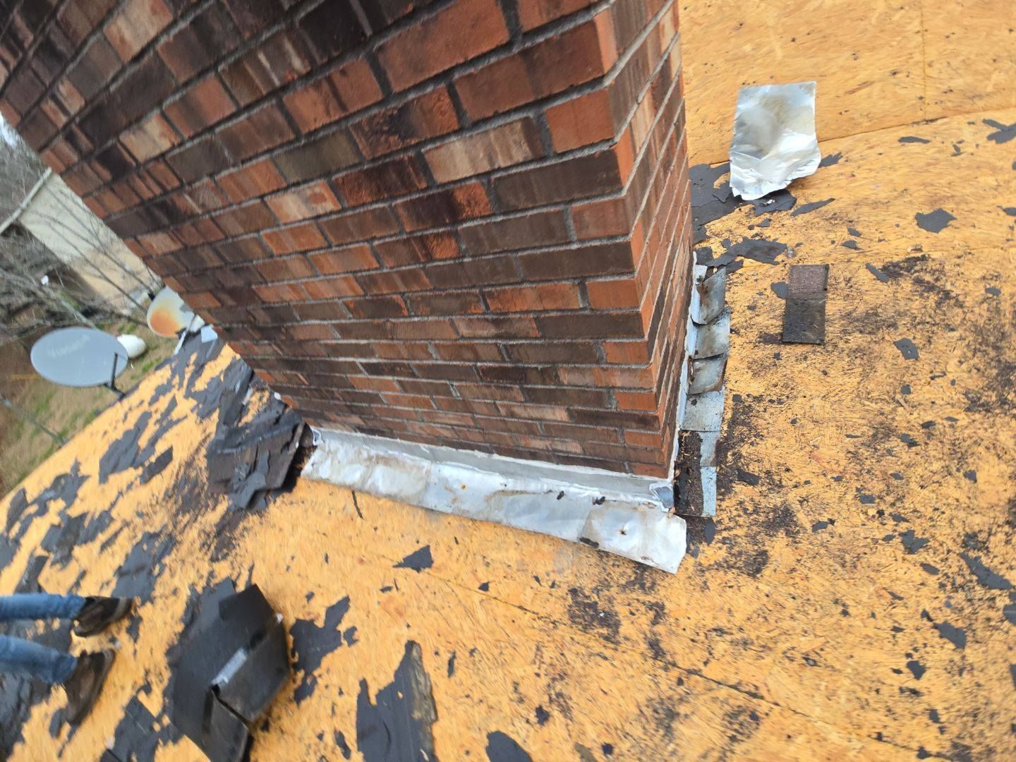Brick chimney on a roof with visible flashing and deteriorating roofing material.