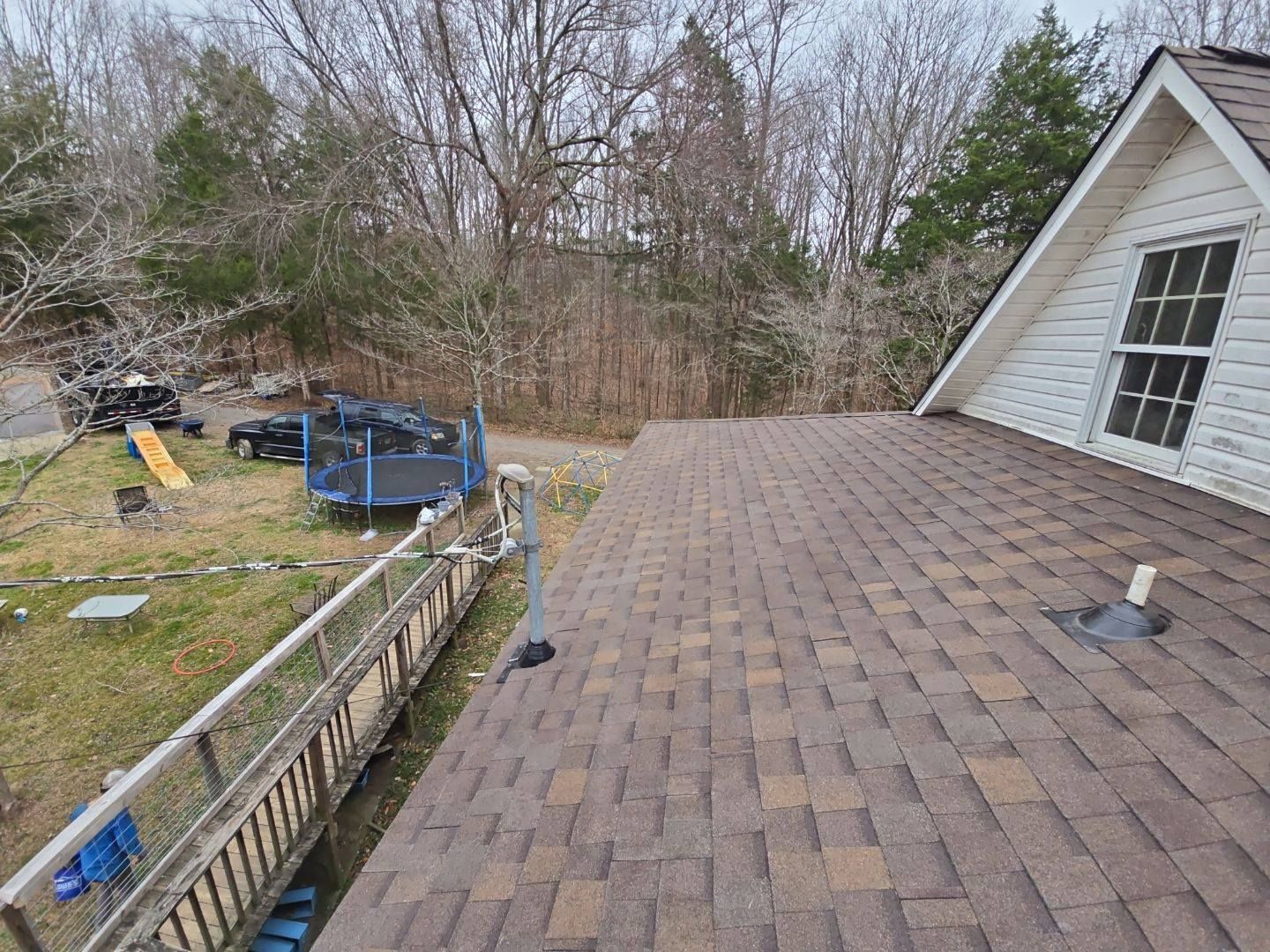 Brown shingle roof of a white house with a gable window, a trampoline, and a wooded background.
