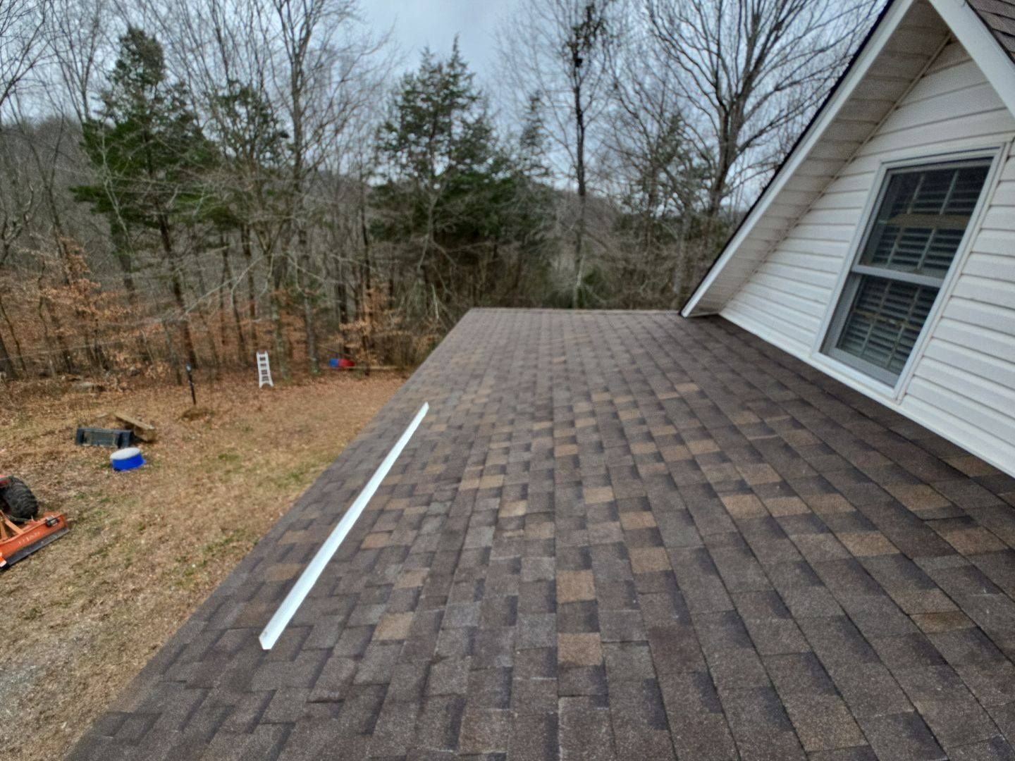 Brown shingled roof of a house with a white trim window and bare trees in the background.