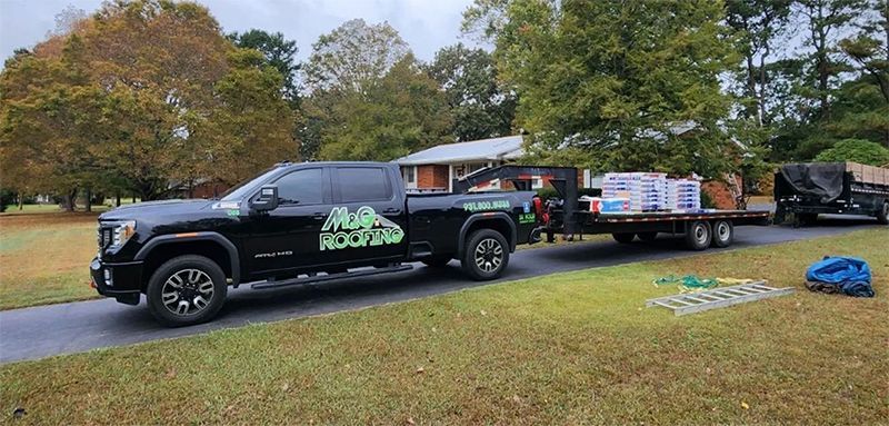 Black truck towing trailers loaded with roofing materials on a driveway. Green lawn, trees, and house in background.