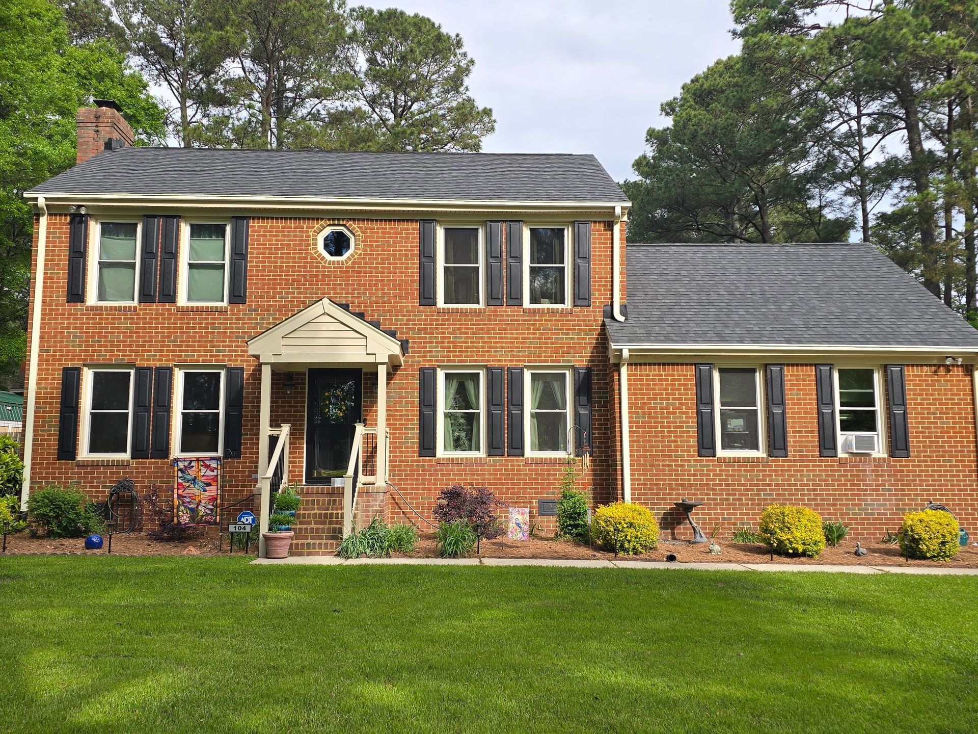 Two-story brick house with green shutters, white porch, and a large lawn under cloudy skies