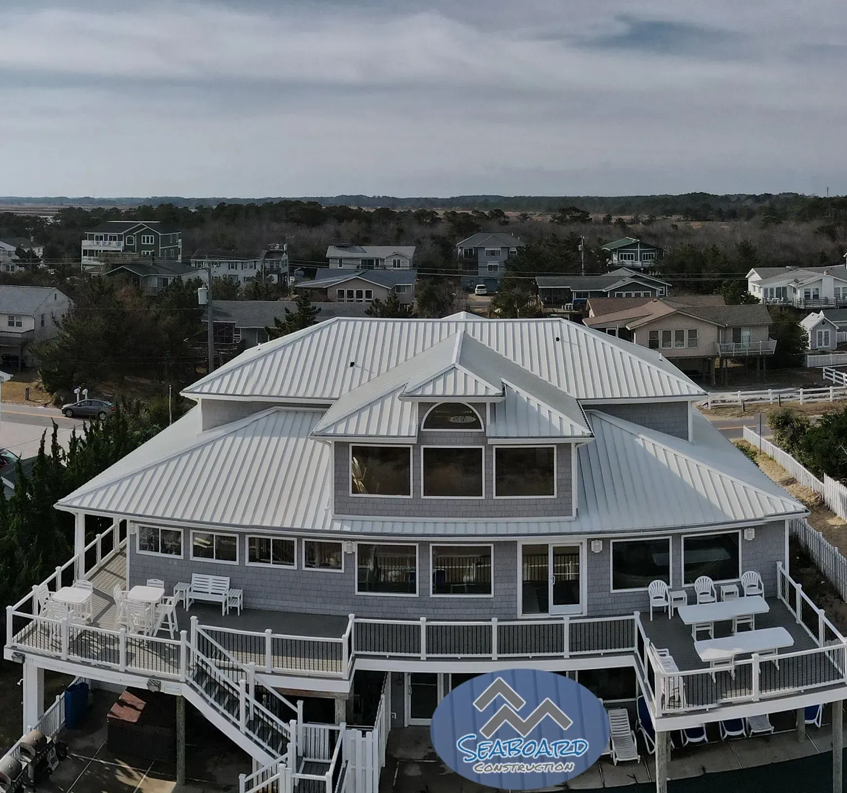 An elevated, light-gray coastal house with a silver metal roof, white railings, and wraparound decks under a cloudy sky.