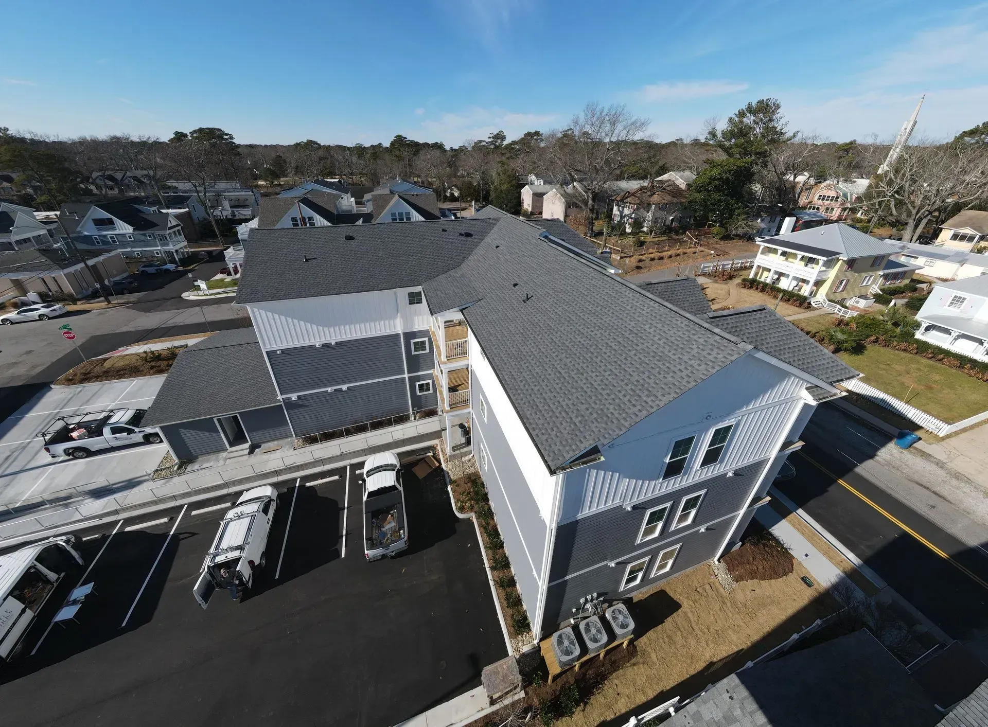 Aerial view of a two-story gray apartment building with a dark shingled roof, parking lot, and surrounding neighborhood.