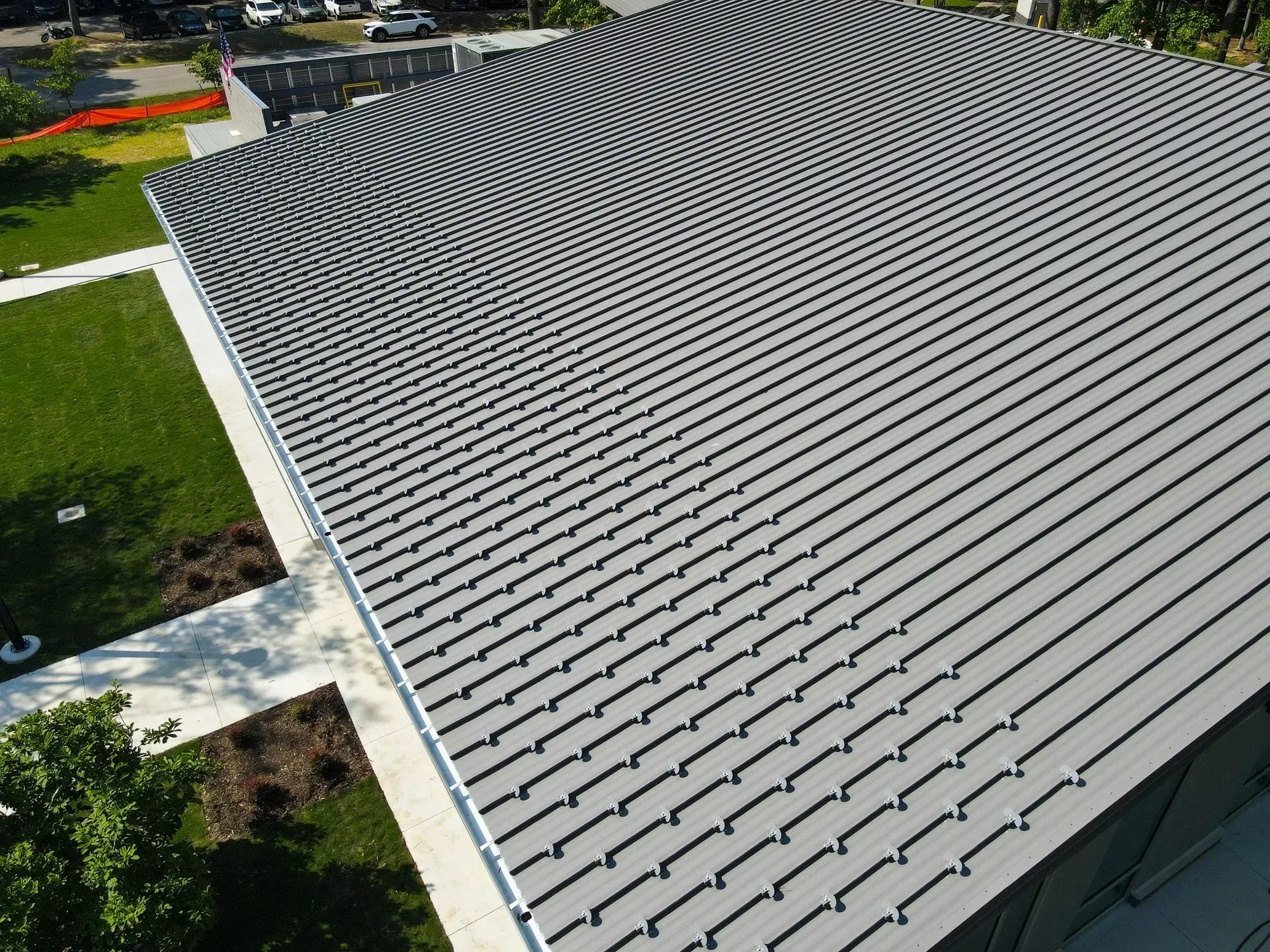 An aerial view of a building with a gray, concave metal roof, next to a landscaped lawn and paved walkway.