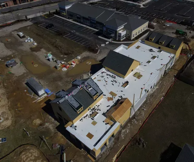 Aerial view of a commercial building under construction with white roofing and surrounding dirt and parking areas.