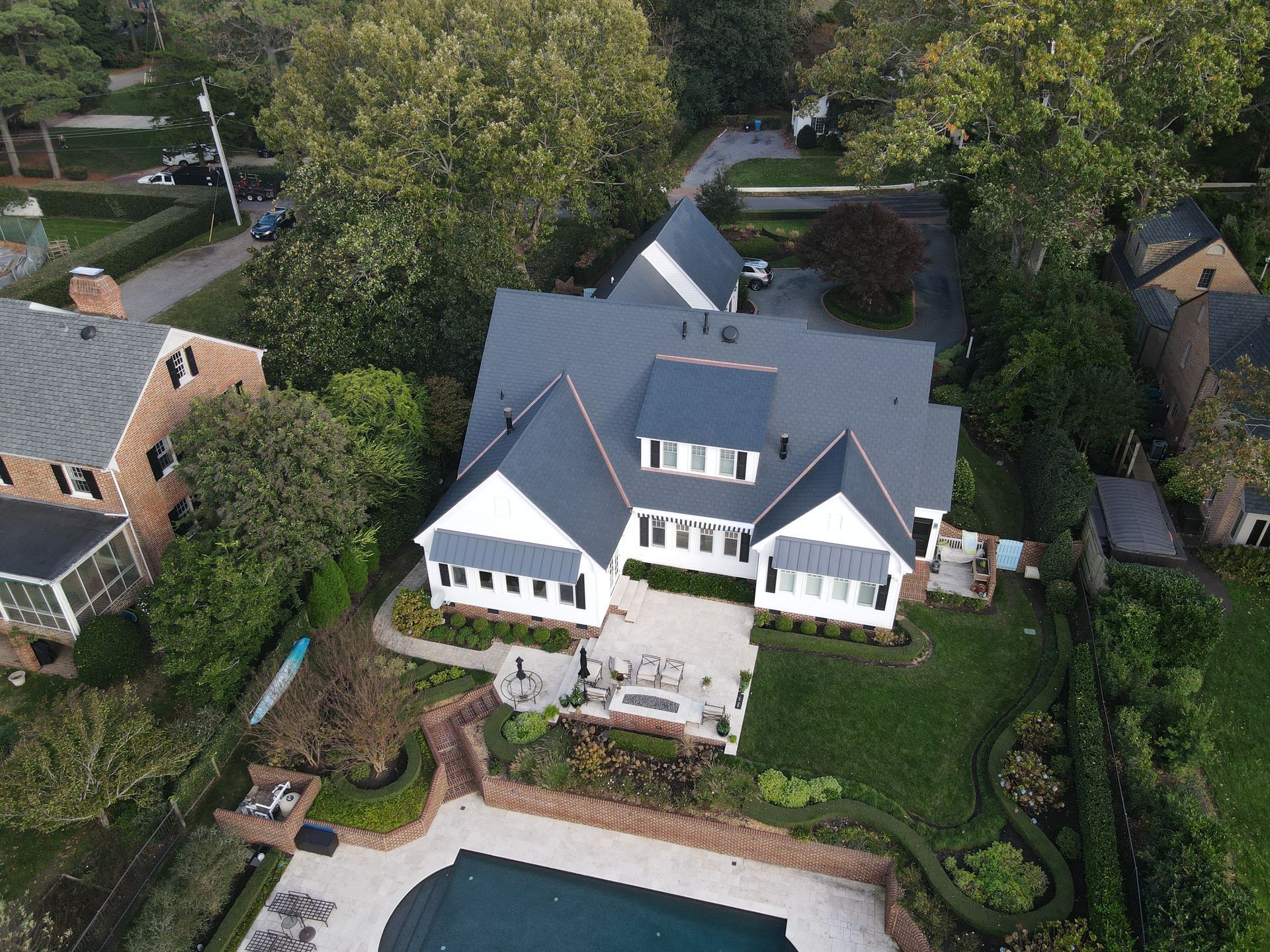 Aerial view of a white, multi-roof house with a blue-gray roof, a back patio, and a swimming pool in a lush yard.