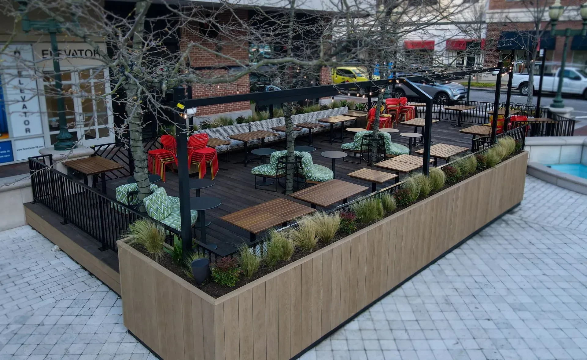An outdoor restaurant patio with wooden planters, tables, and seating, enclosed by a black frame on a tiled plaza.