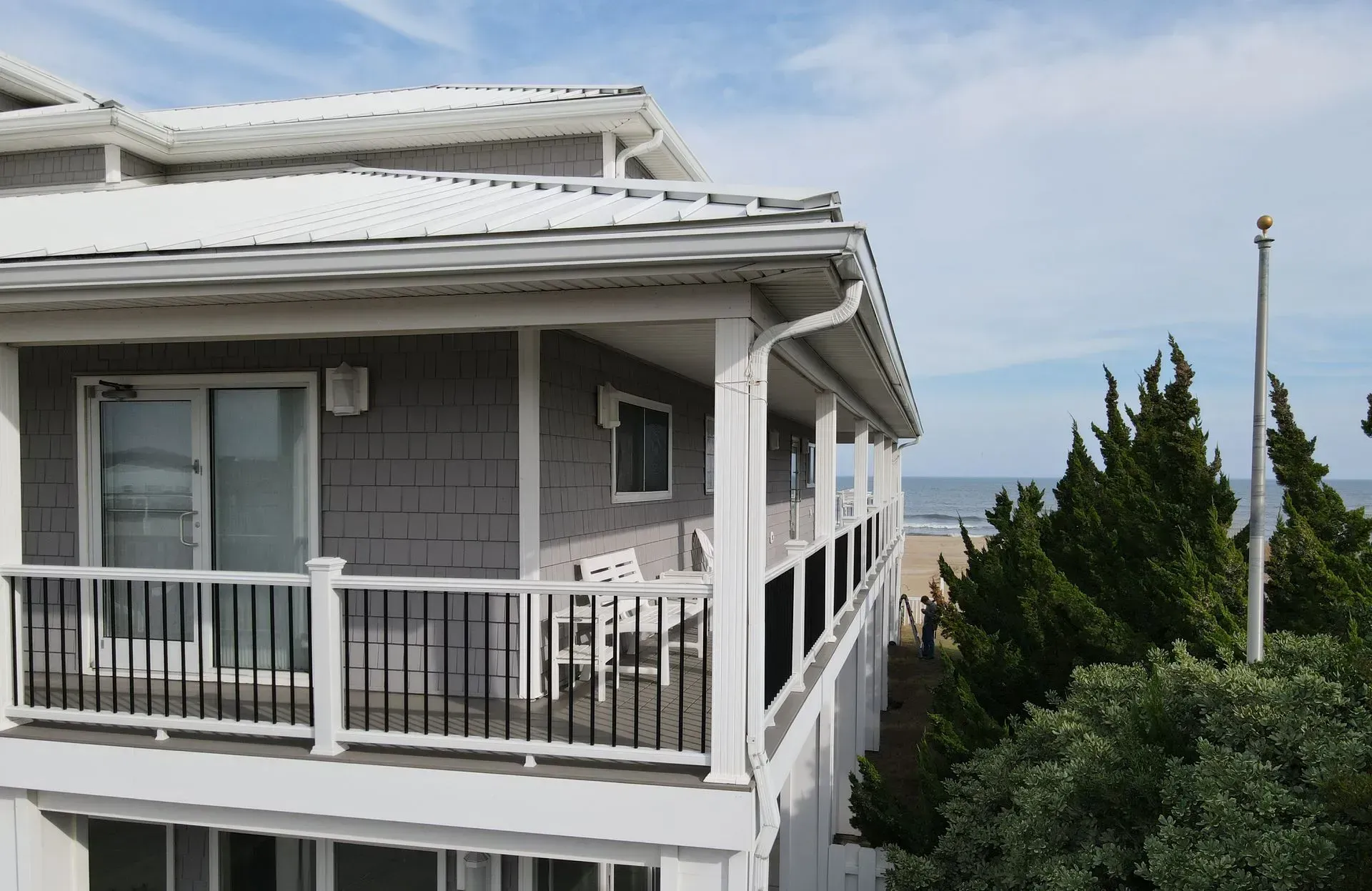 A gray, two-story beachfront house with a white wraparound porch, railing, and roof overlooking the ocean and trees.