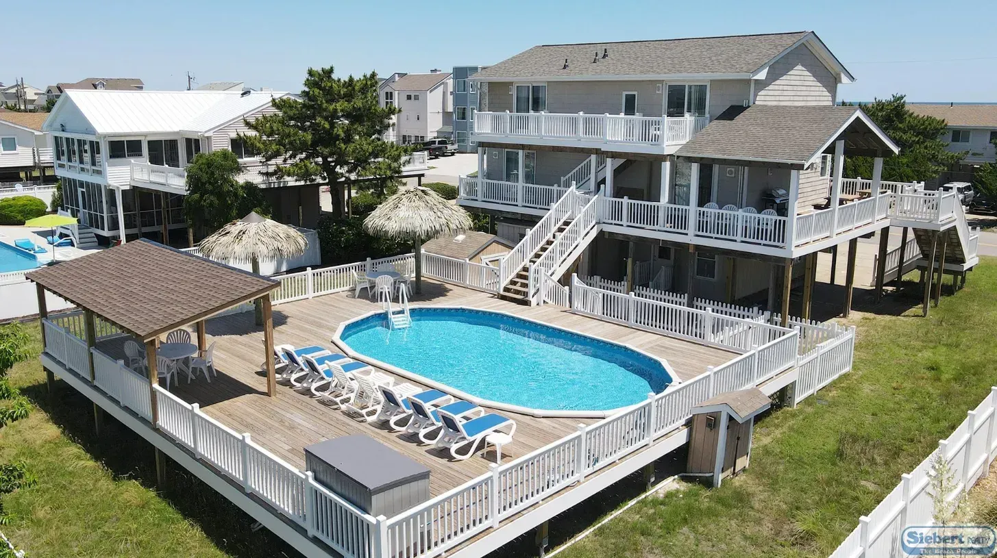 An elevated beach house with a large deck, swimming pool, lounge chairs, and straw umbrellas on a sunny day.