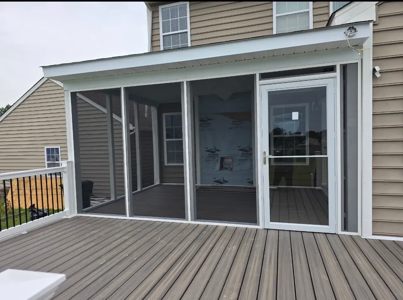 A screened-in porch with white framing and a glass door, situated on a gray composite deck next to a tan house.