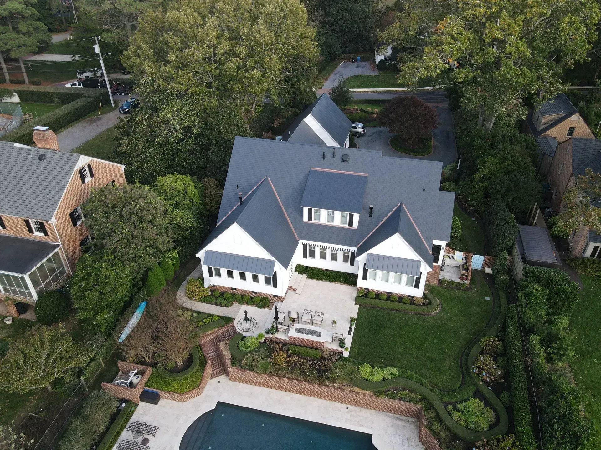 Aerial view of a white, multi-roof house with a pool, brick patio, and manicured landscaping in a suburban neighborhood.
