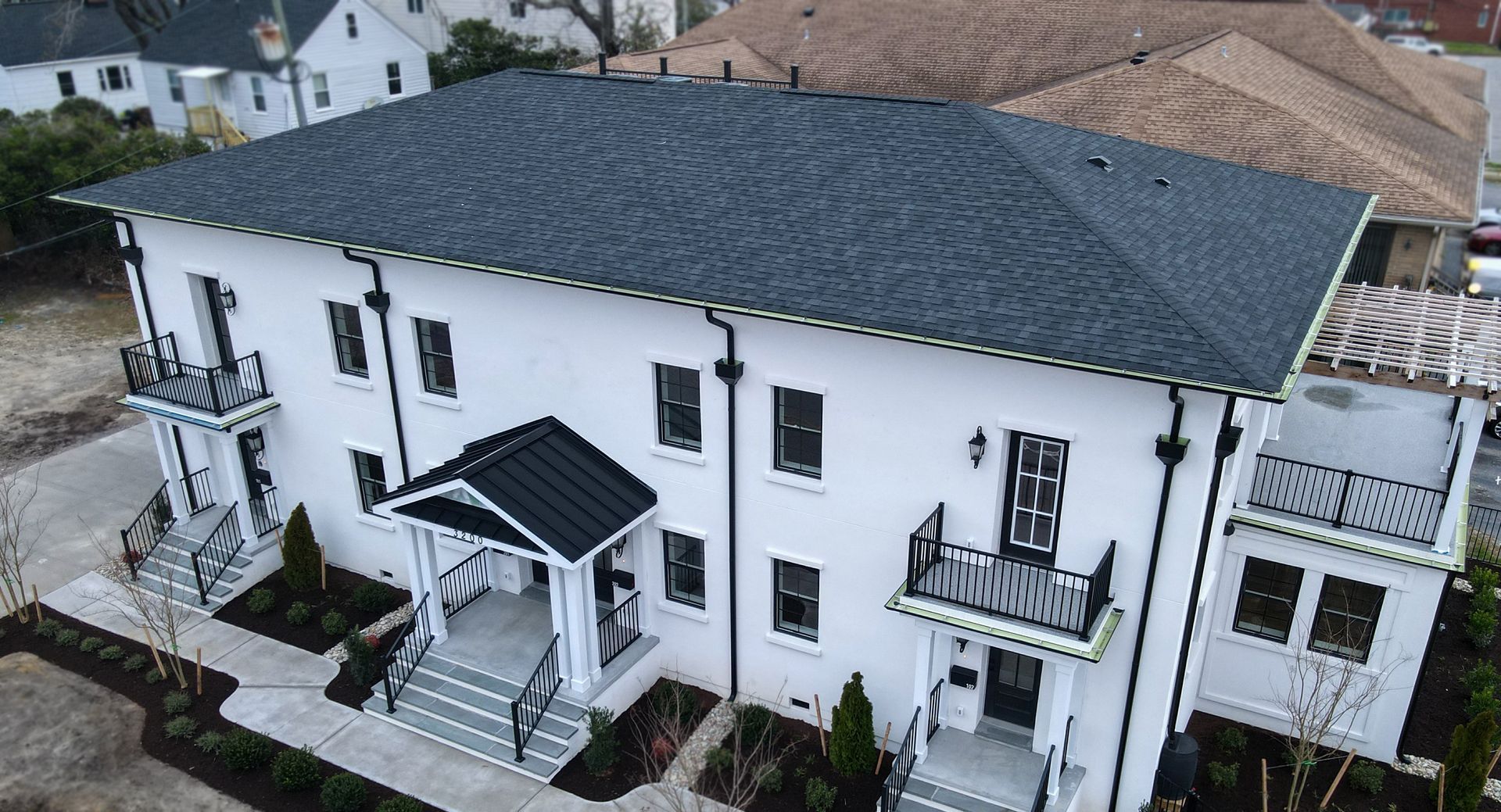 An aerial view of a two-story white building with a dark shingled roof, black accents, balconies, and landscaped grounds.
