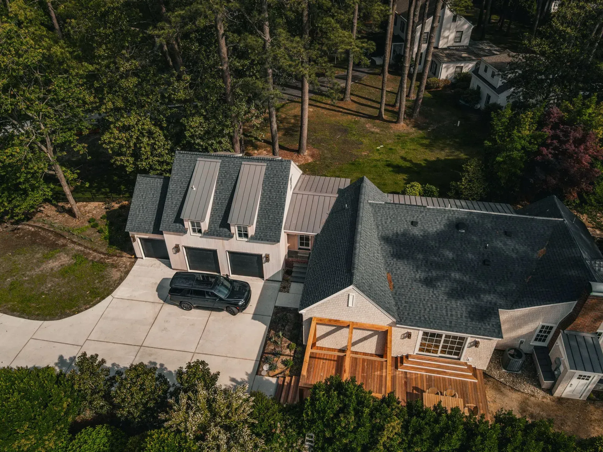 An aerial view of a light-colored house with a dark grey roof and a wooden deck, surrounded by a lush forest and driveway.