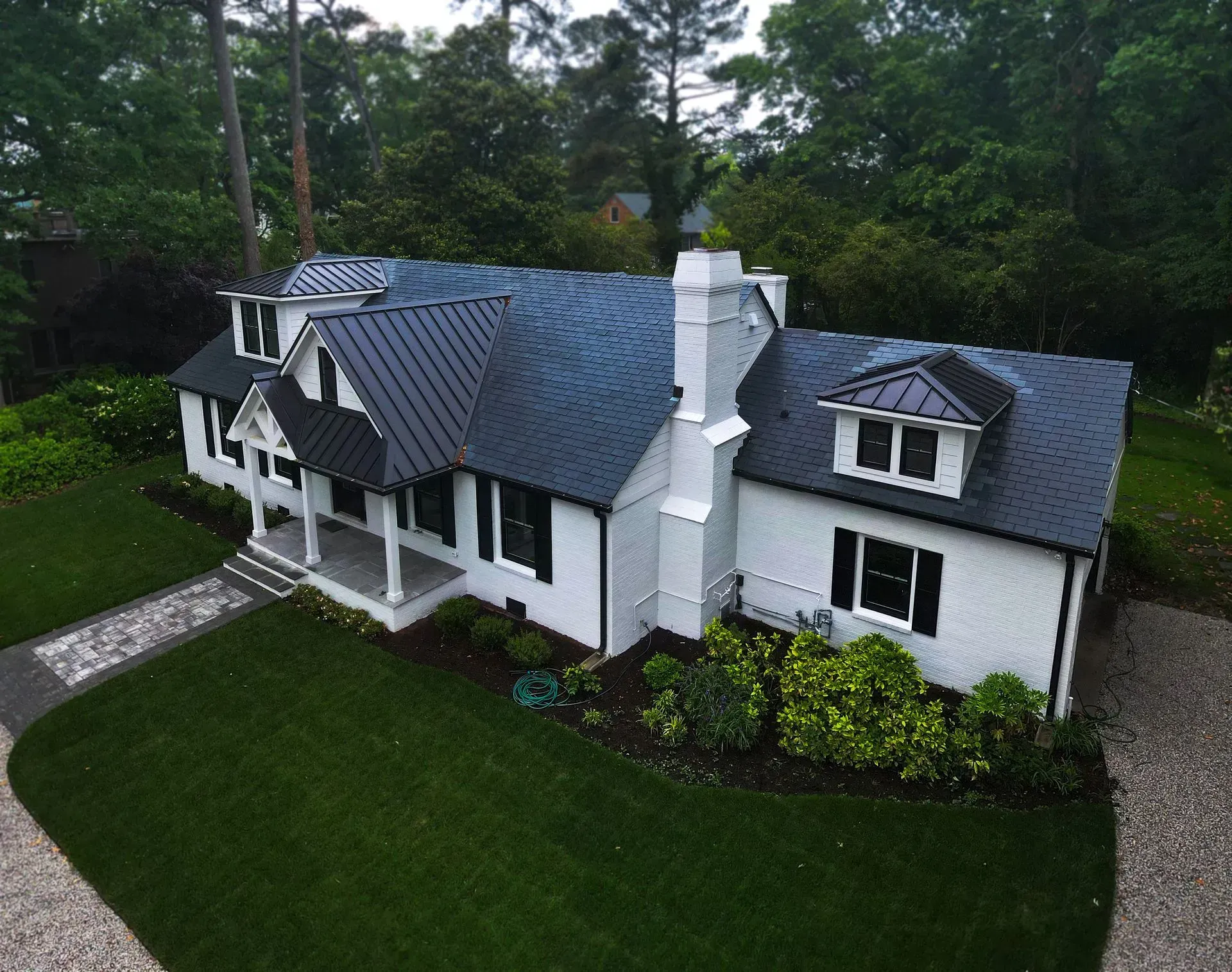 A white brick house with a dark metal roof, black window shutters, a front porch, and a lush green lawn.