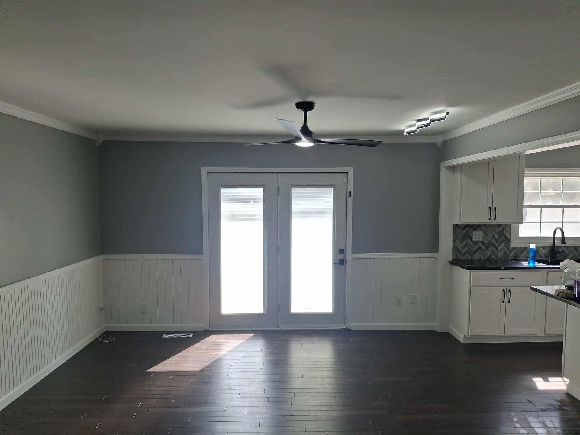 Empty dining room with gray walls, white wainscoting, dark wood floors, and French doors to a bright patio.