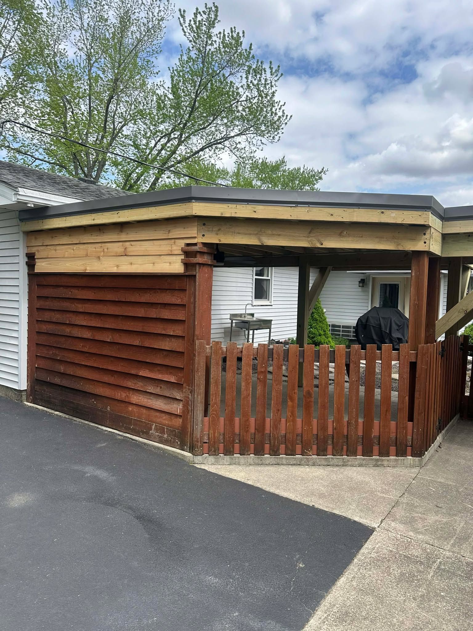 Wooden carport with brown fence and white siding beside a driveway, under a leafy tree.