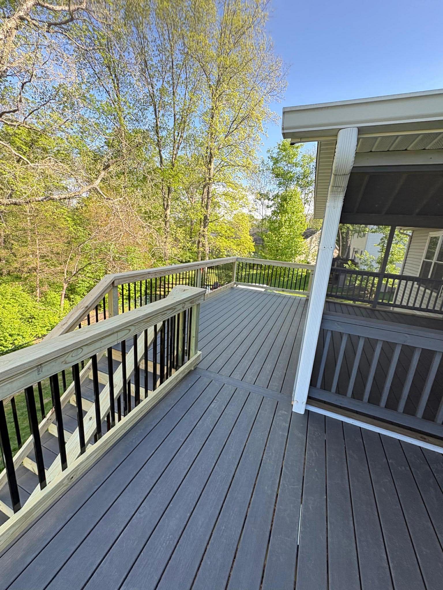 Curved gray deck with black railing beside a house, overlooking green trees on a sunny day