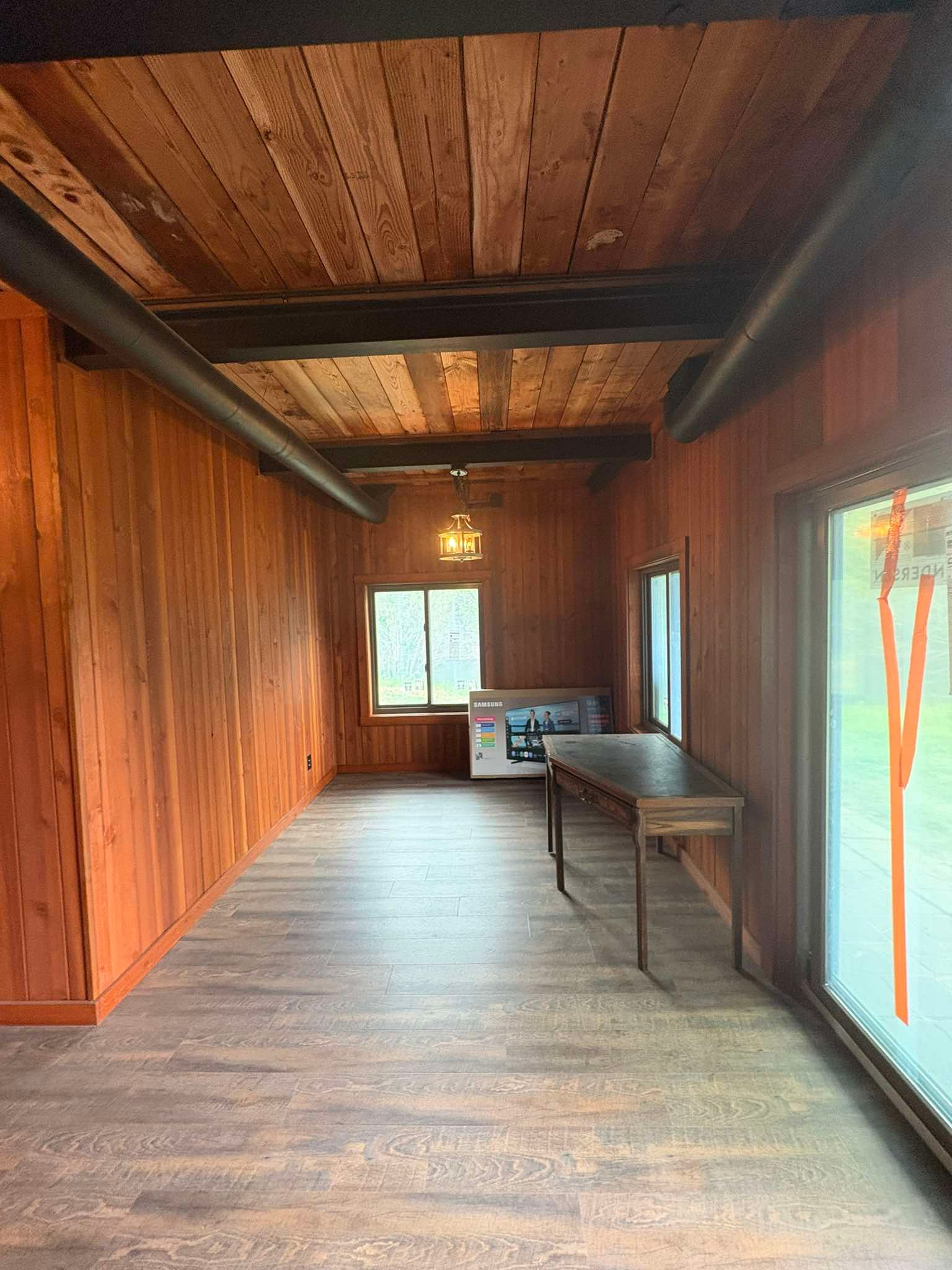 Rustic wood-paneled room with exposed beams, large windows, and a small table at the far end.