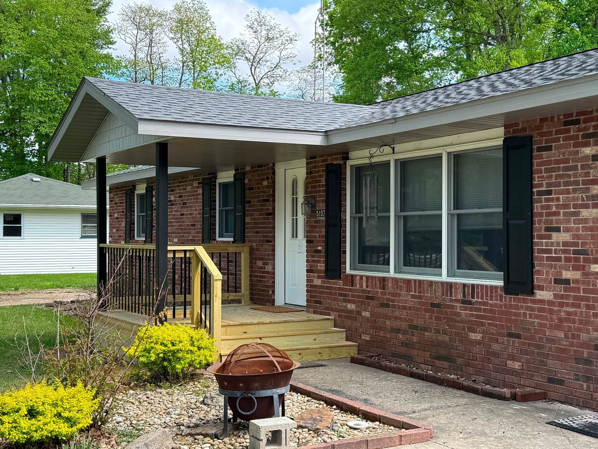 Brick house porch with wooden steps, black railing, and a small rusted fire pit in the front yard