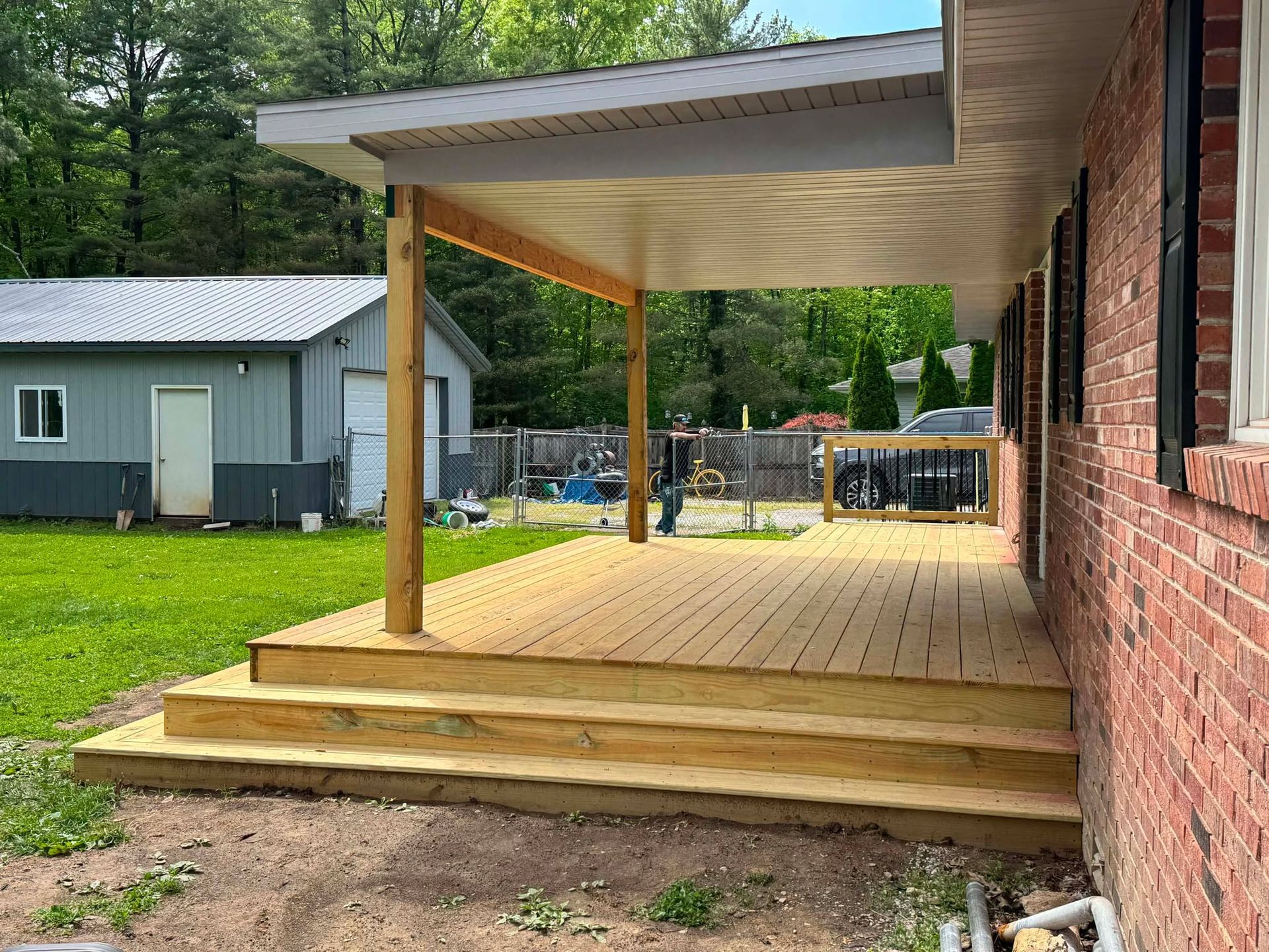 Covered wooden porch beside a brick house, with steps and a metal roof extending over the deck.