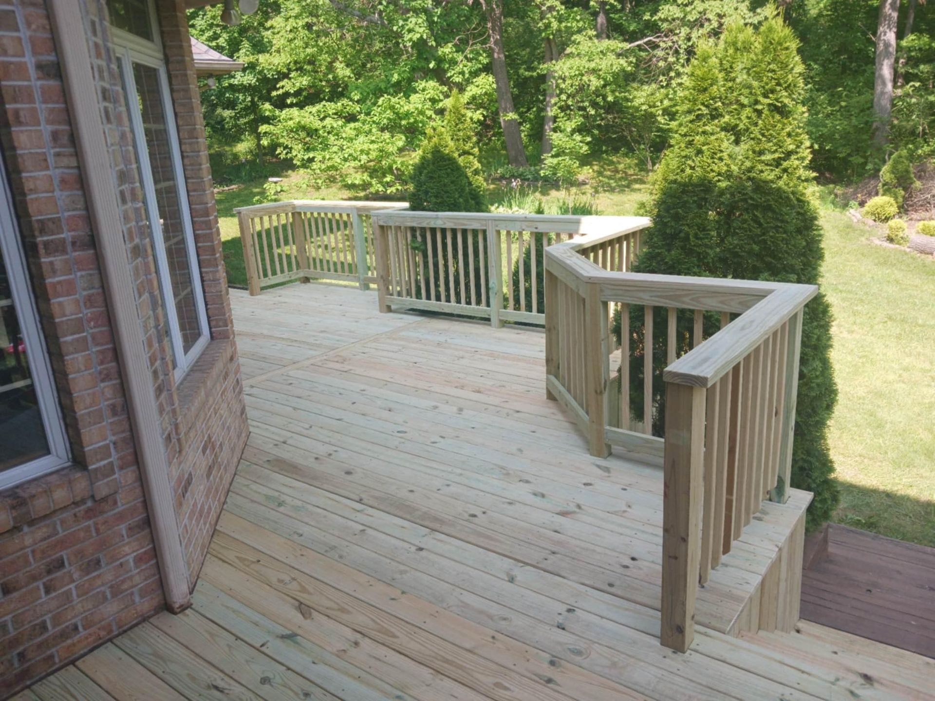 Wooden backyard deck with railings and steps beside a brick house, overlooking a green lawn.