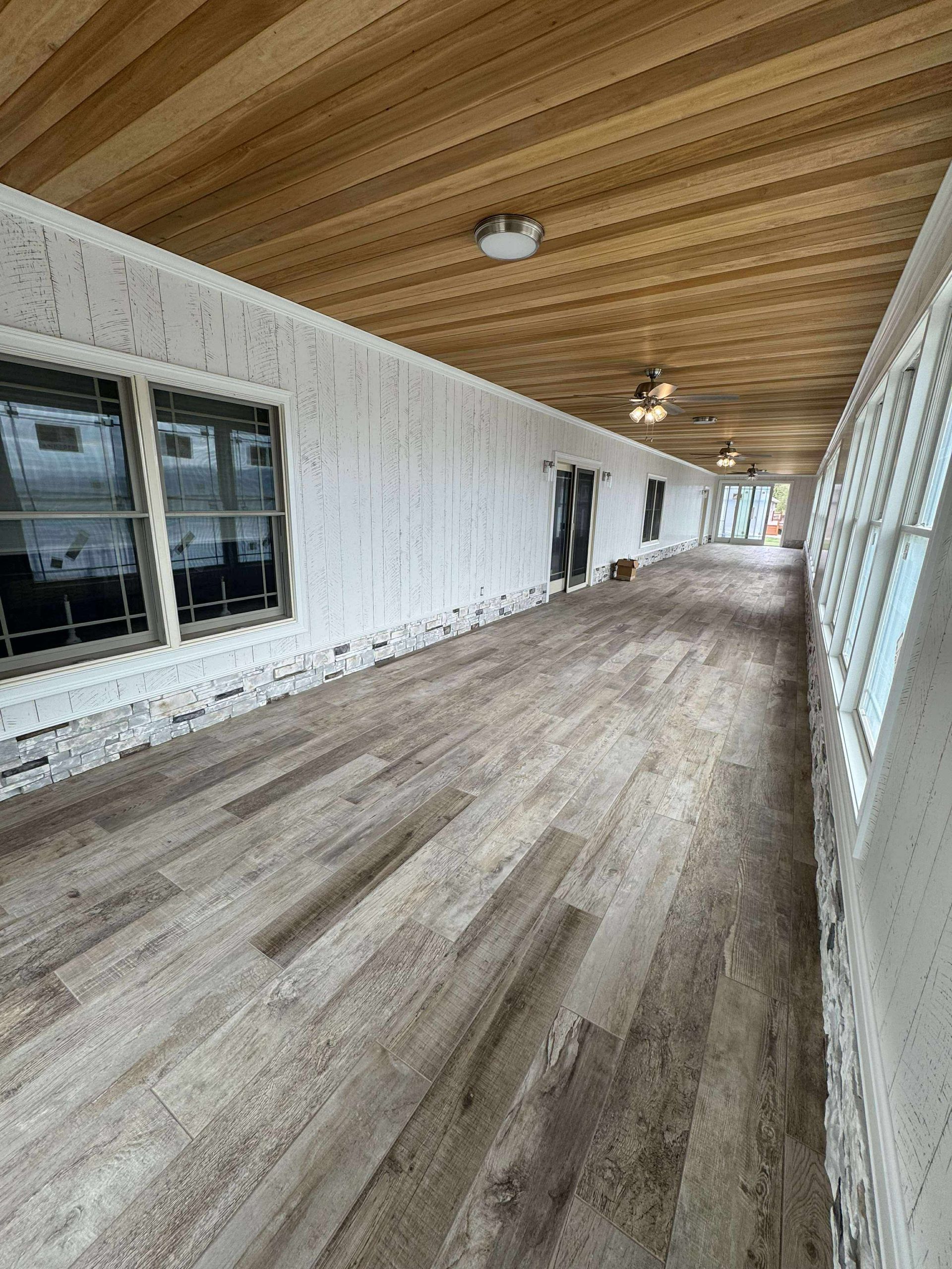 Covered wooden porch with white siding, sloped wood ceiling, and windows along the left wall.