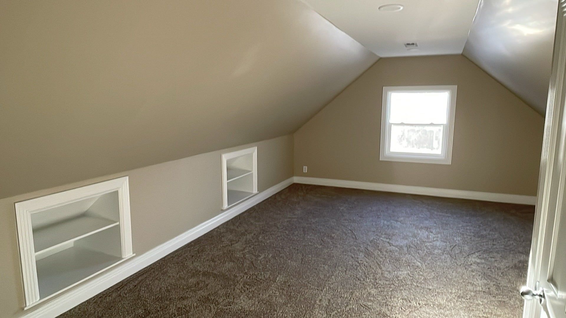 Empty carpeted attic room with sloped beige walls and two small windows.