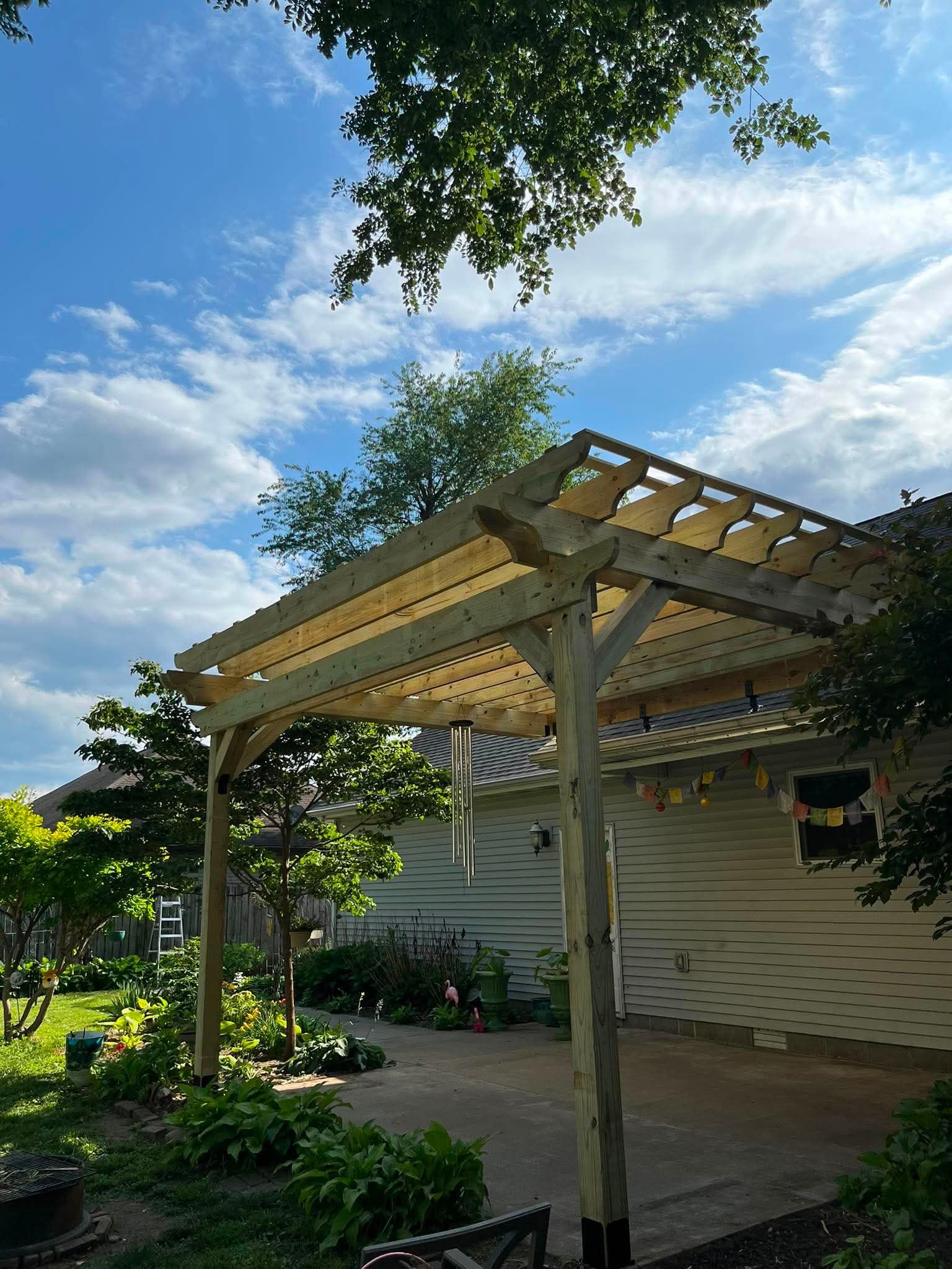 Wooden pergola beside a house with a patio and lush garden under a partly cloudy sky