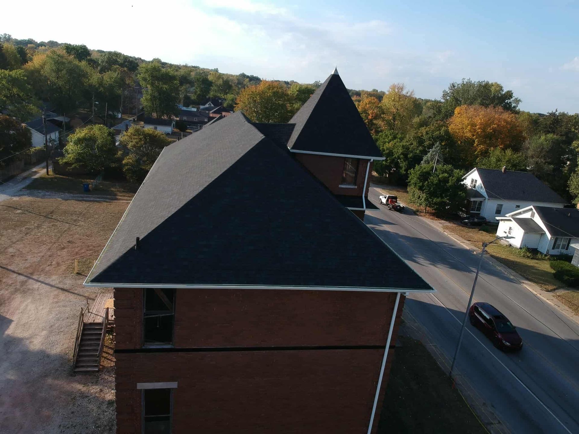 Aerial view of a brick building with a dark roof beside a road and parked car, surrounded by trees