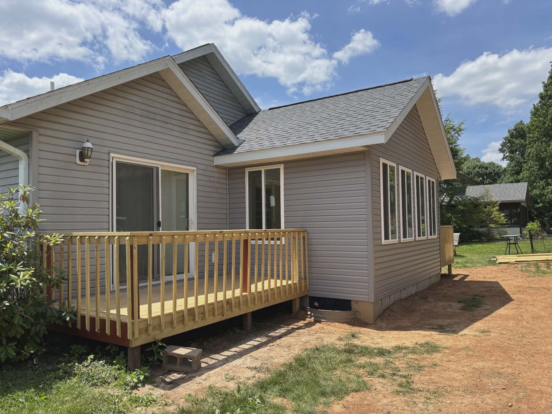 Small gray house with a wooden front deck and steps in a dirt yard under a partly cloudy sky