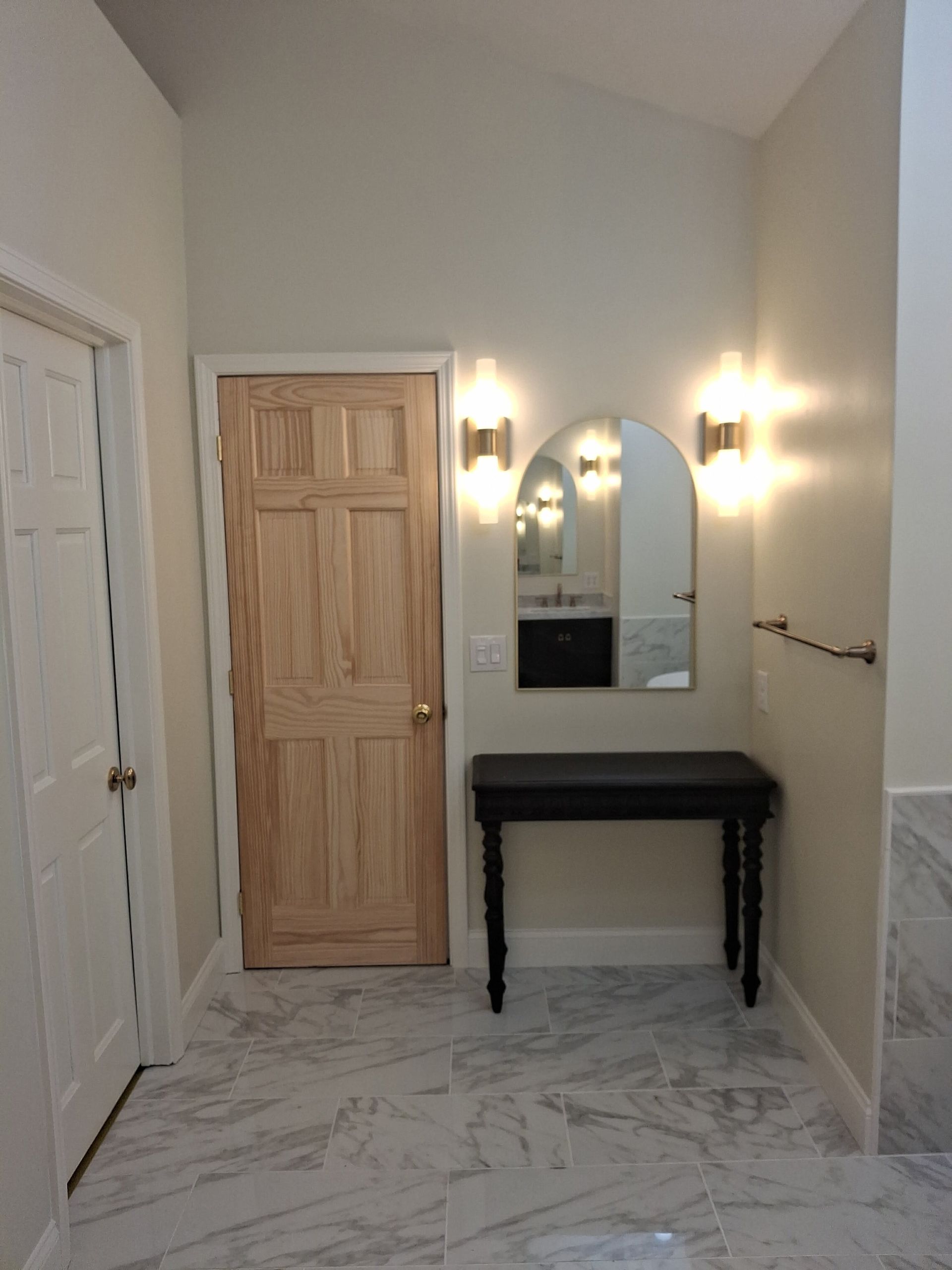 Hallway with marble floor, black console table, mirror, wall sconces, and wooden door