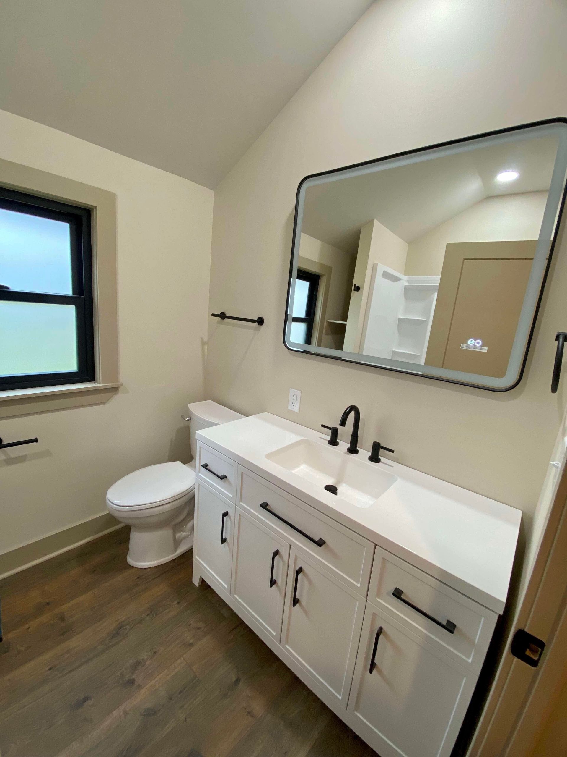 Modern bathroom with white vanity, black fixtures, mirror, toilet, and wood-look floor.