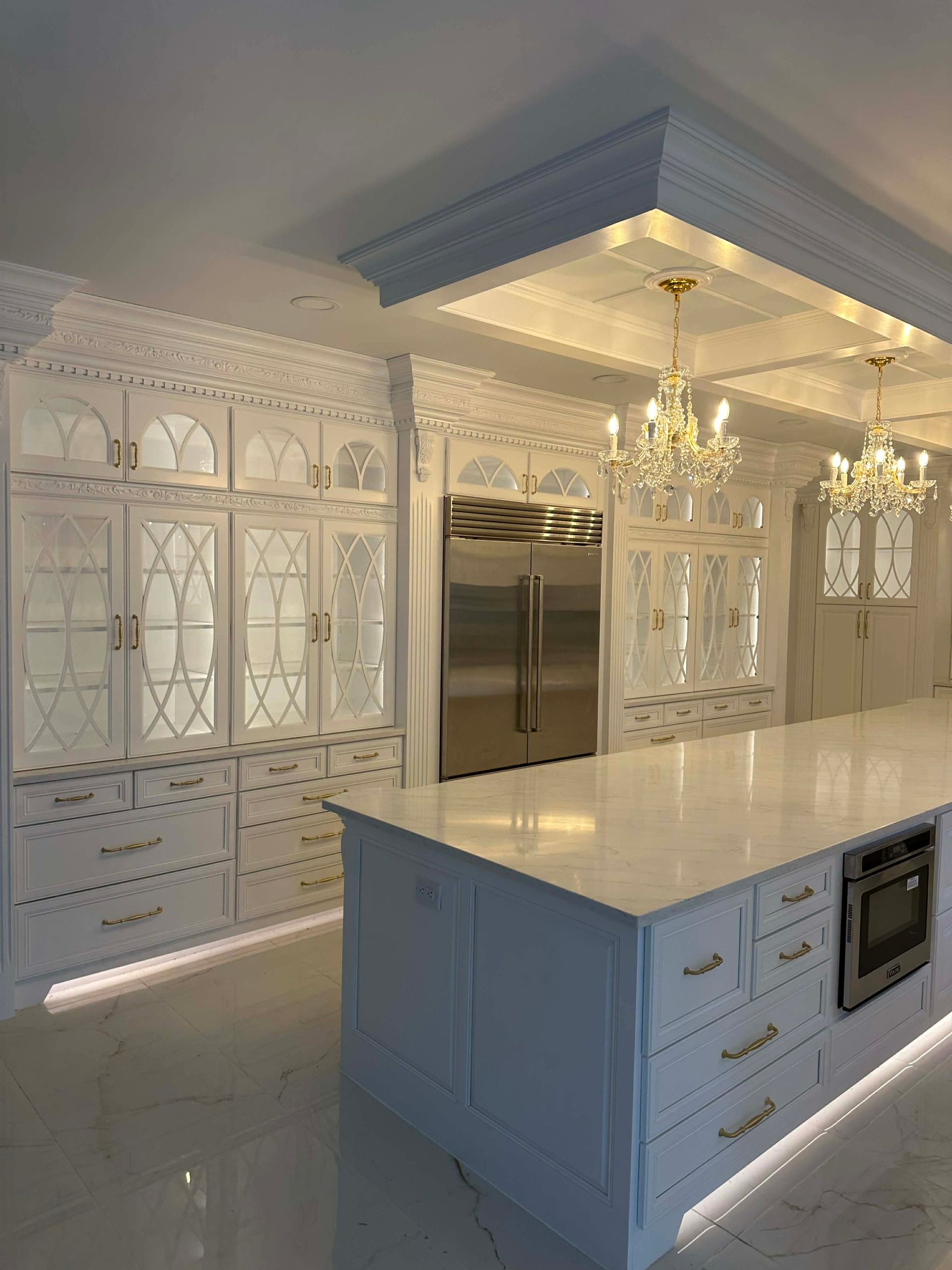 Bright white ornate kitchen with large island, stainless steel fridge, and decorative ceiling lights.