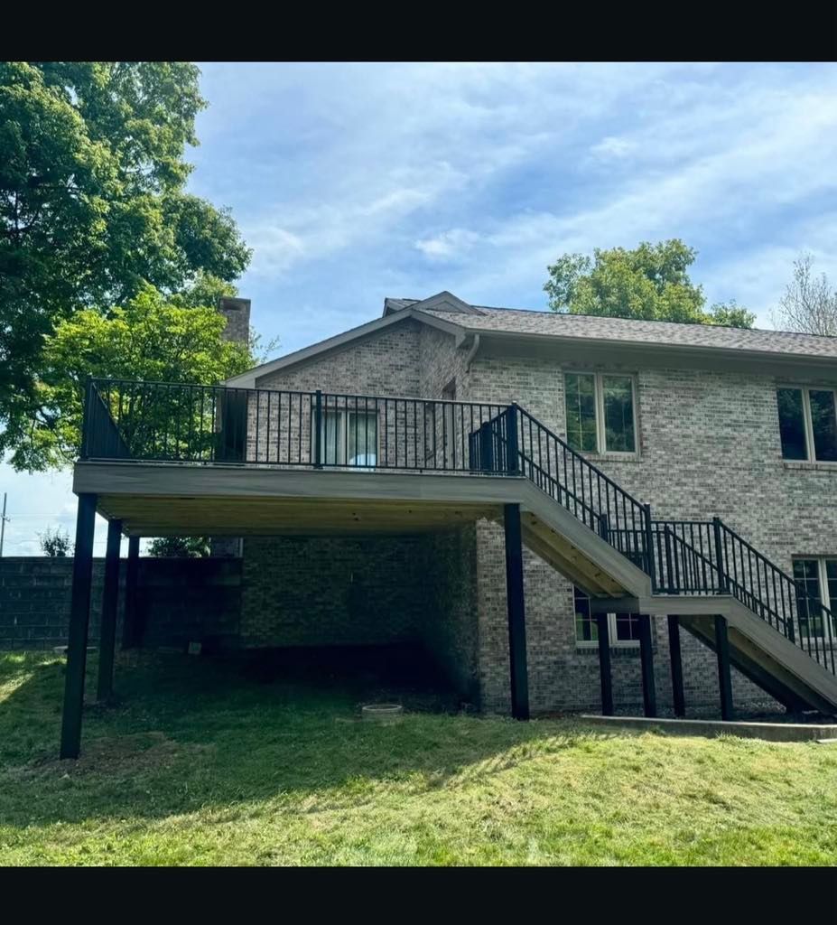 Stone house with a raised deck and exterior staircase, set in a grassy yard with trees.