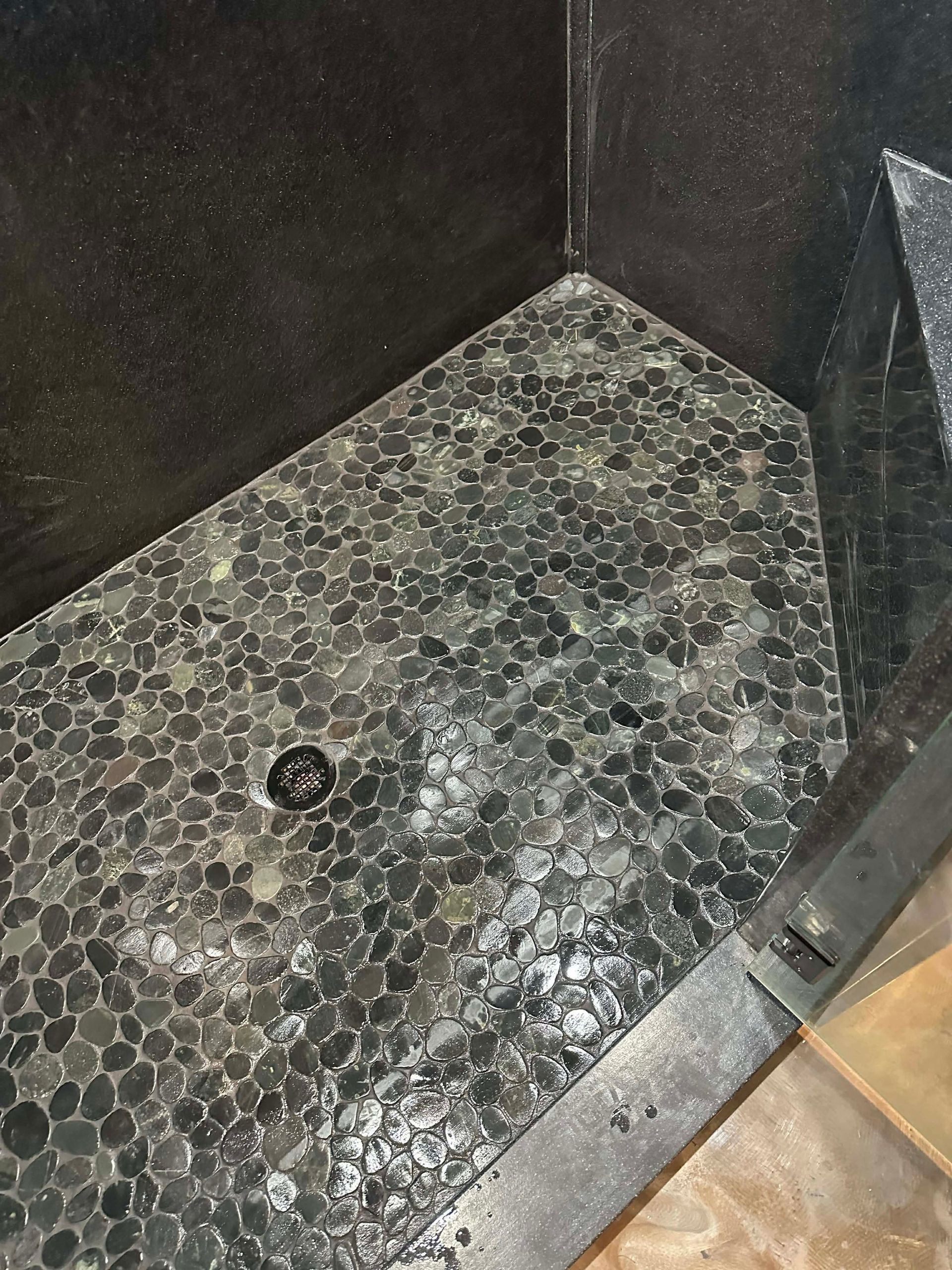 Small corner shower floor with pebble mosaic tiles and a drain, bordered by dark walls.